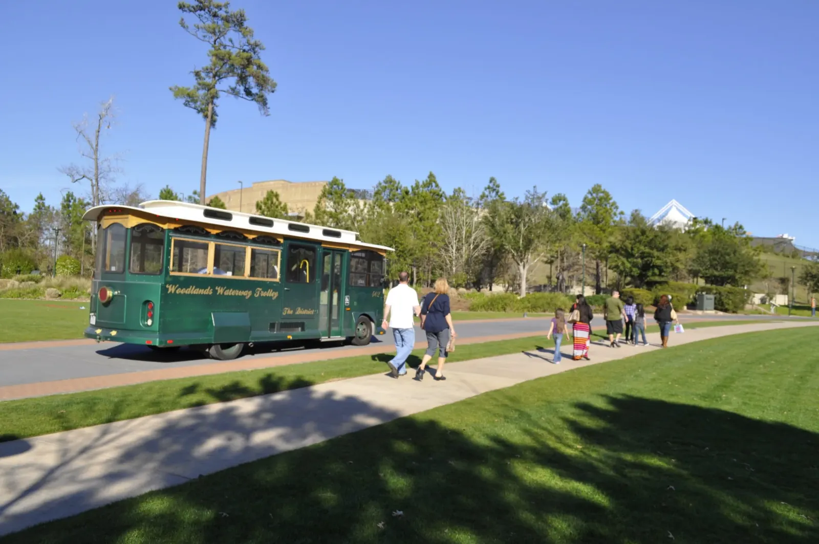 Green Woodlands Waterway Trolley on the Waterway in Town Center, with walkers on the path and The Cynthia Woods Mitchell Pavilion behind the trees